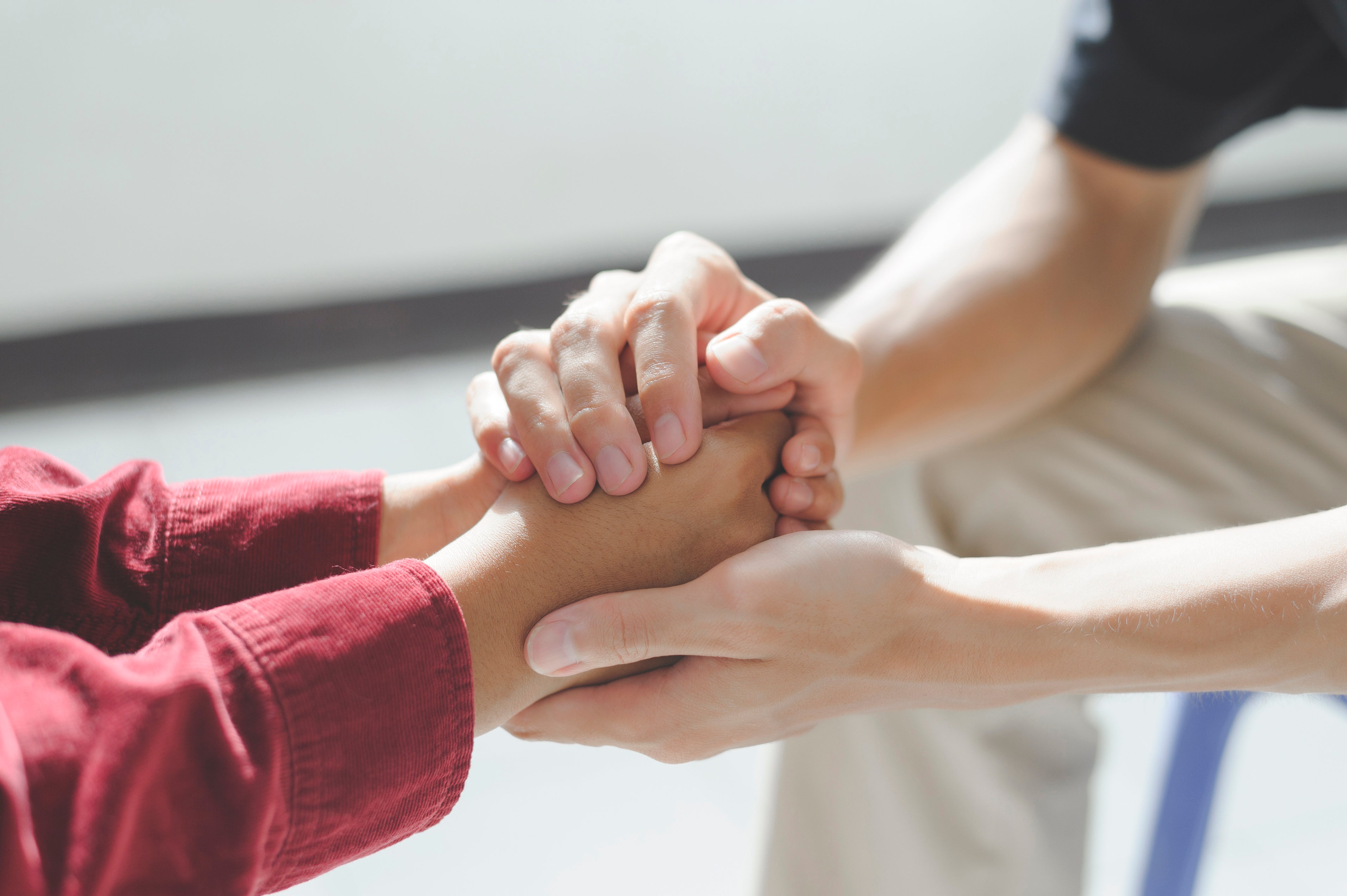 Eine Nahaufnahme zeigt eine mitfühlende Geste: Eine Person umschließt tröstend die Hände einer anderen Person, die ein rotes Oberteil trägt.