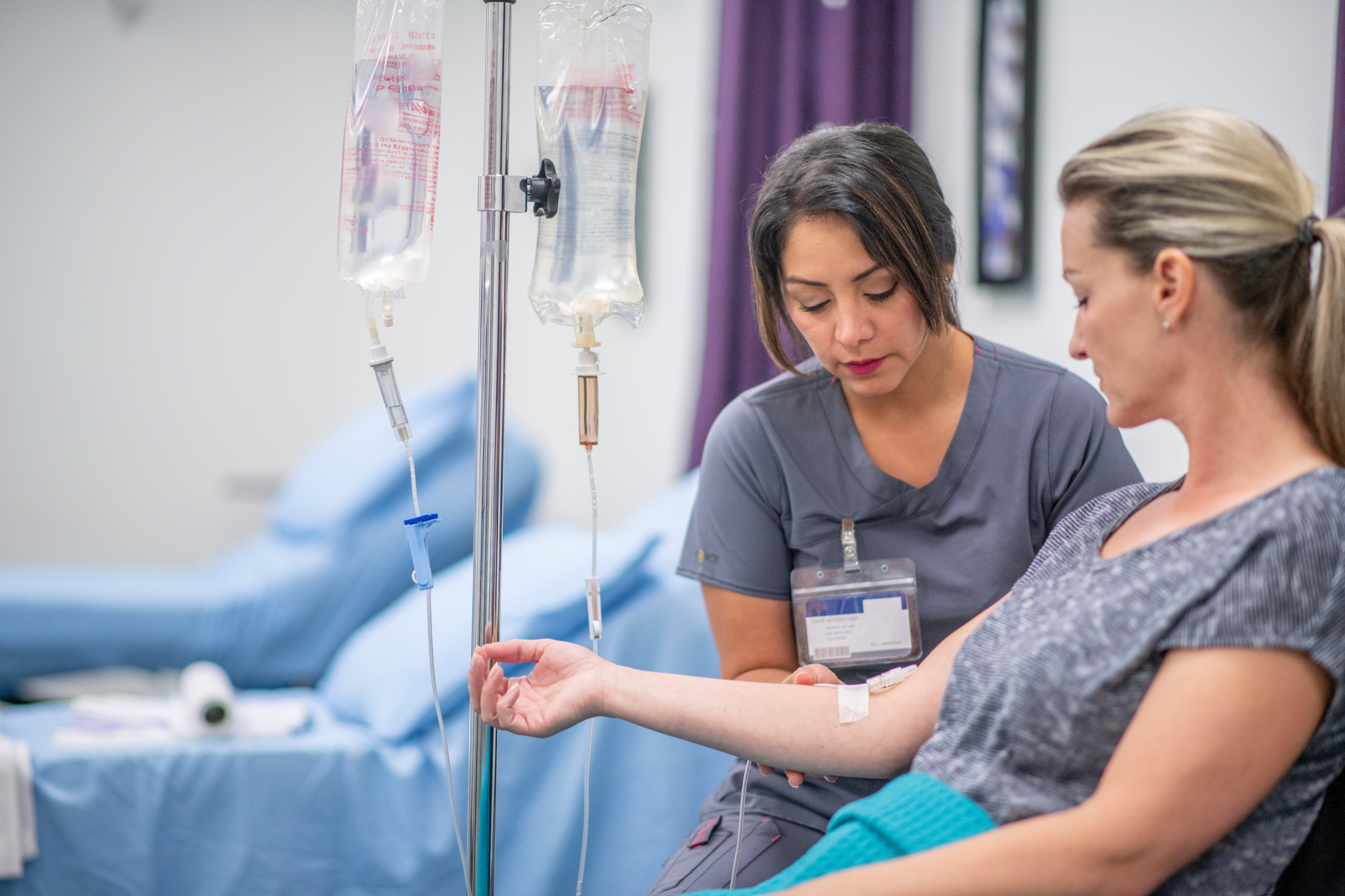 Eine medizinische Fachkraft in grauer Arbeitskleidung kümmert sich in einem Behandlungsraum konzentriert um eine sitzende Patientin.