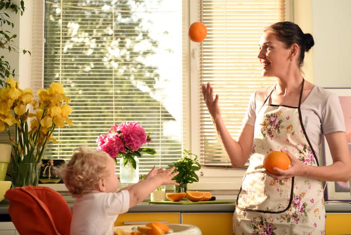 In einer sonnendurchfluteten Küche wirft eine lächelnde Frau mit einer geblümten Schürze spielerisch eine Orange in die Luft.