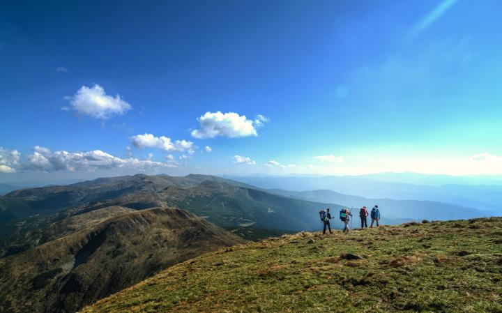 Eine weitwinklige Landschaftsaufnahme zeigt eine Gruppe von vier Wanderern mit großen Rucksäcken von hinten, die auf einem grasbewachsenen Bergrücken nach rechts gehen.