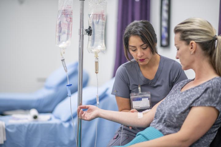 Eine medizinische Fachkraft in grauer Arbeitskleidung kümmert sich in einem Behandlungsraum konzentriert um eine sitzende Patientin.
